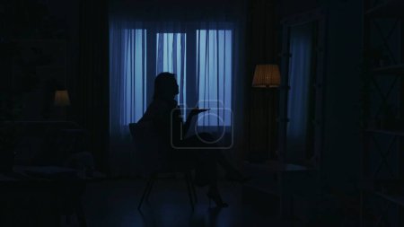 Téléchargez les photos : Portrait d'une silhouette féminine dans la pièce sombre de l'appartement. La vie quotidienne concept créatif. Femme dans une robe assise sur une chaise dans la chambre parlant sur smartphone. - en image libre de droit
