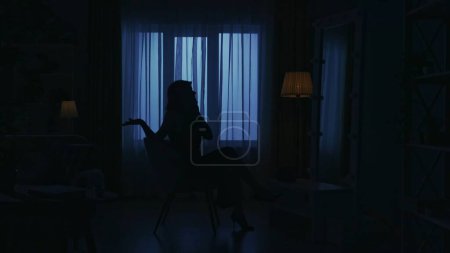 Téléchargez les photos : Portrait d'une silhouette féminine dans la pièce sombre de l'appartement. La vie quotidienne concept créatif. Femme en robe assise sur une chaise dans le salon et parlant sur smartphone. - en image libre de droit