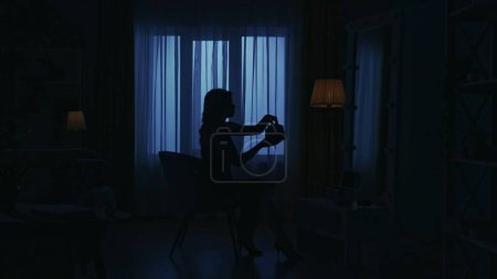 Photo for Portrait of female silhouette in the dark apartment. Everyday life creative concept. Woman in a dress sitting on a chair in the room, looking in the mirror, holding cosmetic purse. - Royalty Free Image