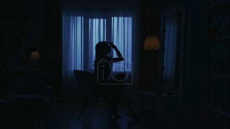 Téléchargez les photos : Portrait de silhouette féminine dans l'appartement sombre. La vie quotidienne concept créatif. Femme dans une robe assise sur une chaise dans la chambre, regardant dans le miroir, fixant son maquillage. - en image libre de droit