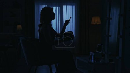 Photo for Portrait of female silhouette in the dark apartment. Everyday life creative concept. WCloseup shot. Woman in a dress sitting on a chair in the room, looking in the mirror, fixing makeup with a brush. - Royalty Free Image