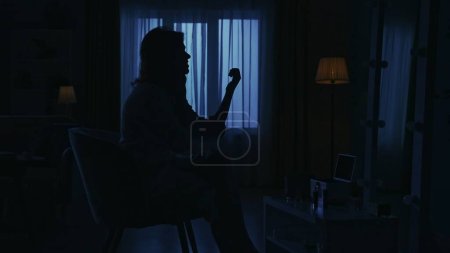 Photo for Portrait of female silhouette in the dark apartment. Everyday life creative concept. Closeup shot. Woman in bathrobe sitting on a chair in the room, looking at her nails, talking on the phone. - Royalty Free Image