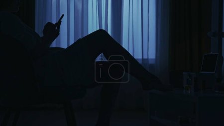 Photo for Portrait of female silhouette in the dark apartment. Everyday life creative concept. Closeup shot. Woman in bathrobe sitting on a chair in the living room, holding smartphone. - Royalty Free Image