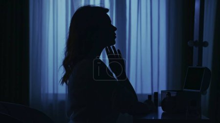Téléchargez les photos : Portrait de silhouette féminine dans l'appartement sombre. La vie quotidienne concept créatif. Femme en peignoir assise sur une chaise dans la chambre, regardant dans le miroir, touchant son visage. - en image libre de droit