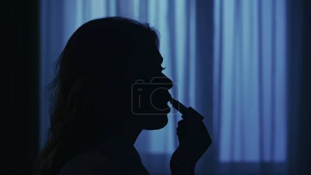 Téléchargez les photos : Portrait de silhouette féminine dans l'appartement sombre. La vie quotidienne concept créatif. Femme en peignoir assise sur une chaise, regardant dans le miroir, appliquant du rouge à lèvres. Gros plan du visage. - en image libre de droit