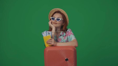 Téléchargez les photos : Publicité itinérante concept créatif. Femme voyageuse en chapeau de paille et lunettes avec valise tenant les billets d'avion, souriant en levant les yeux. Isolé sur fond vert. - en image libre de droit
