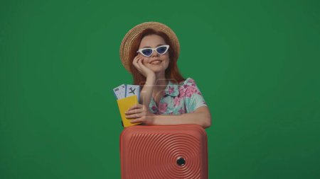 Téléchargez les photos : Publicité itinérante concept créatif. Femme voyageuse en chapeau de paille et lunettes avec valise tenant les billets d'avion, souriant en levant les yeux et rêvant. Isolé sur fond vert. - en image libre de droit