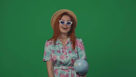 Foto de Concepto creativo de publicidad itinerante. Mujer viajera con sombrero de paja y gafas sosteniendo un globo mirando a la cámara feliz expresión de la cara. Aislado sobre fondo verde. - Imagen libre de derechos