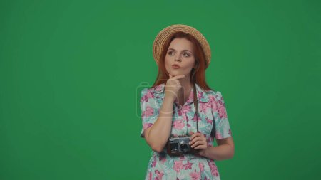 Téléchargez les photos : Publicité itinérante concept créatif. Femme en chapeau de paille tenant la caméra de film, pensant à quelque chose avec le visage concentré. Isolé sur fond vert. - en image libre de droit