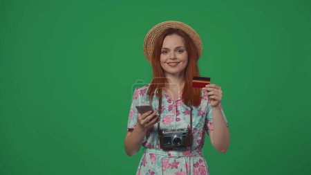 Photo for Travelling advertisement creative concept. Woman traveller in straw hat holding credit card and smartphone, online payments service, smiling face expression. Isolated on green background. - Royalty Free Image
