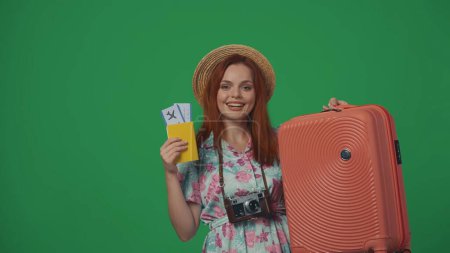 Téléchargez les photos : Publicité itinérante concept créatif. Femme voyageuse en chapeau de paille tenant billets d'avion, passeport et valise, souriant expression joyeuse du visage. Isolé sur fond vert. - en image libre de droit