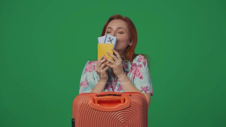 Téléchargez les photos : Publicité itinérante concept créatif. Voyageuse avec valise contenant des billets d'avion, expression joyeuse et souriante du visage, embrassant les billets. Isolé sur fond vert. - en image libre de droit
