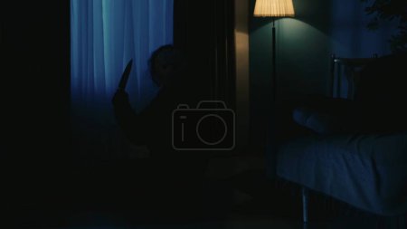 Photo for Horror movie scene. Halloween advertisement concept. Closeup shot of man maniac silhouette sitting on the floor in the dark apartment room near window and holding knife in hand. - Royalty Free Image