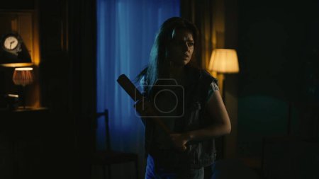 Photo for Horror movie scene. Halloween advertisement concept. Young girl with scaried face expression standing in the dark living room, looking around, holding baseball bat. - Royalty Free Image