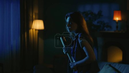 Photo for Horror movie scene. Halloween advertisement concept. Young girl with scaried face expression walking in the dark living room, looking around, holding baseball bat in hand. - Royalty Free Image