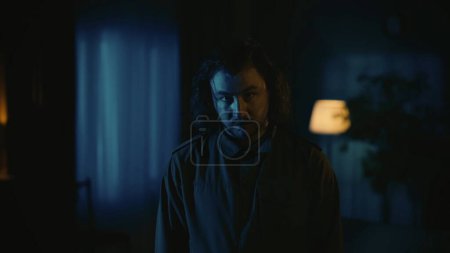 Photo for Horror movie scene. Halloween advertisement concept. Man maniac with long hair standing in the dark apartment room, looking directly at the camera with straight face, tense atmosphere. - Royalty Free Image