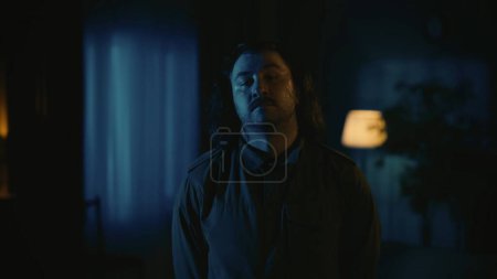 Photo for Horror movie scene. Halloween advertisement concept. Man maniac with long hair standing in the dark living room, looking directly at the camera, posing with head up. - Royalty Free Image