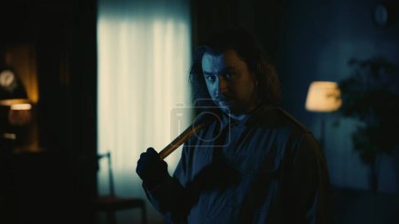 Photo for Horror movie scene. Halloween advertisement concept. Man maniac with long hair standing in the dark living room, looking at the camera and holding baseball bat. - Royalty Free Image