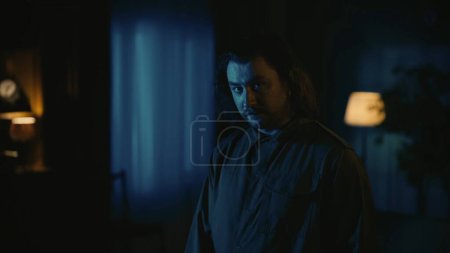 Photo for Horror movie scene. Halloween advertisement concept. Man maniac with long hair standing in the dark apartment living room, looking at the camera with serious face expression. - Royalty Free Image