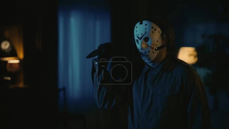 Photo for Horror movie scene. Halloween advertisement concept. Man maniac in hockey mask standing in the someones dark apartment, holding knife in hand, posing at hte camera, terrified atmosphere. - Royalty Free Image