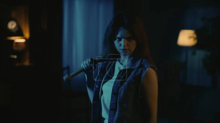 Photo for Horror movie scene. Halloween advertisement concept. Girl in casual clothing posing with baseball bat standing in the living room, looking at the camera with serious face expression. - Royalty Free Image