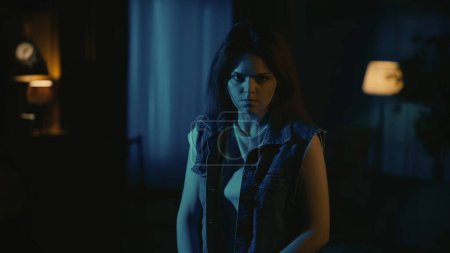 Photo for Horror movie scene. Halloween advertisement concept. Girl in casual clothing with angry face expression looking at the camera standing in the dark living room, tense atmosphere. - Royalty Free Image