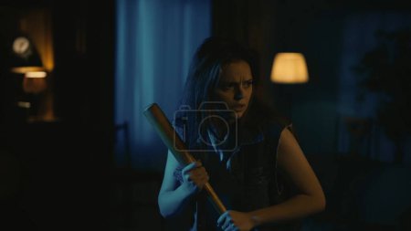 Photo for Horror movie scene. Halloween advertisement concept. Young girl with scared face holding baseball bat and looking around for maniac, nervous atmosphere, thunderstorm outside. - Royalty Free Image