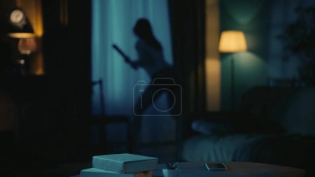 Photo for Horror movie scene. Halloween advertisement concept. Afraid young girl with baseball bat standing near the window in the dark living room, looking outside. - Royalty Free Image