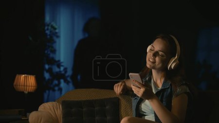 Photo for Horror movie scene. Halloween advertisement concept. Girl in headphones sitting on the sofa in the living room, listening to music, man maniac standing with knife near window. - Royalty Free Image