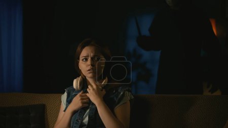 Photo for Horror movie scene. Halloween advertisement concept. Girl in headphones sitting on the sofa in the dark living room with scaried face, man maniac standig behind her back with knife. - Royalty Free Image