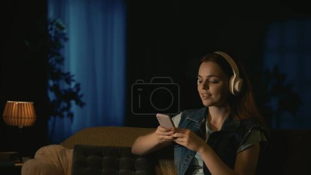 Foto de Escena de película de terror. Concepto de anuncio de Halloween. Chica en auriculares sentados en el sofá en la sala de estar oscura, sin preocupaciones escuchando música en el teléfono inteligente, expresión de la cara sonriente. - Imagen libre de derechos