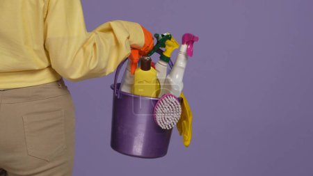Photo for Everyday cleaning and housekeeping concept. Closeup shot of woman in rubber gloves holding bucket with different cleansers, rags, brushes and sponges. Isolated on purple background. - Royalty Free Image