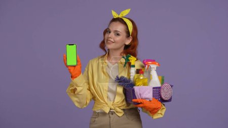 Photo for Everyday cleaning and housekeeping concept. Woman in casual clothing and rubber gloves holding smartphone and basket with cleansers and rugs. Advertising area, workspace mockup. - Royalty Free Image