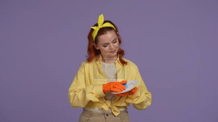 Photo for Everyday cleaning and housekeeping concept. Woman in casual clothing and rubber gloves holding plate and sponge, wiping it and cleaning. Isolated on purple background. - Royalty Free Image