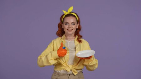 Photo for Everyday cleaning and housekeeping concept. Woman in casual clothing and rubber gloves holding plate and sponge, shows thumbs up smiling at the camera. Isolated on purple background. - Royalty Free Image