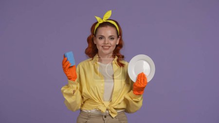 Photo for Everyday cleaning and housekeeping concept. Woman in casual clothing and rubber gloves holding plate and sponge, smiling at the camera. Isolated on purple background. - Royalty Free Image
