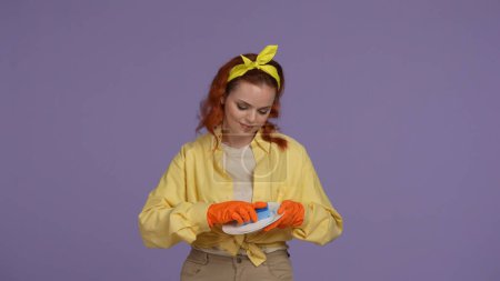 Photo for Everyday cleaning and housekeeping concept. Woman in casual clothing and rubber gloves holding plate and cleaning it with sponge. Isolated on purple background. - Royalty Free Image
