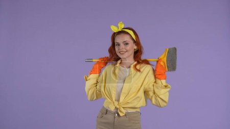 Photo for Everyday cleaning and housekeeping concept. Woman in casual clothing and rubber gloves holding brush on shoulders, looking at the camera. Isolated on purple background. - Royalty Free Image