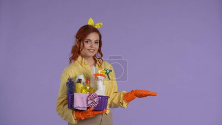 Photo for Everyday cleaning and housekeeping concept. Woman in casual clothing and rubber gloves holding basket with cleaners, showing the empty area with hand. Isolated on purple background. - Royalty Free Image