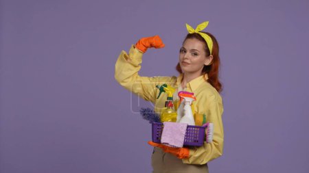 Photo for Everyday cleaning and housekeeping concept. Woman in casual clothing and rubber gloves with basket of cleaning tools, holding hand up and smiling. Isolated on purple background. - Royalty Free Image