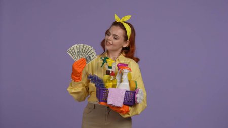 Photo for Everyday cleaning and housekeeping concept. Woman in casual clothing and rubber gloves holding fan of money and basket of cleansers and rugs, looking them. Isolated on purple background. - Royalty Free Image