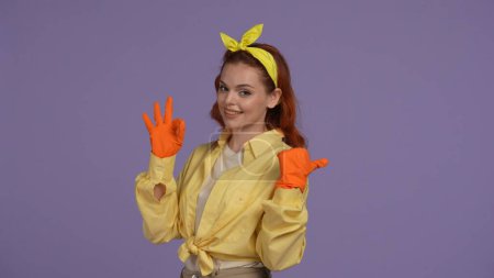 Photo for Everyday cleaning and housekeeping concept. Woman in casual clothing and rubber gloves showing ok sign pointing at the empty area, happy face. Isolated on purple background. - Royalty Free Image