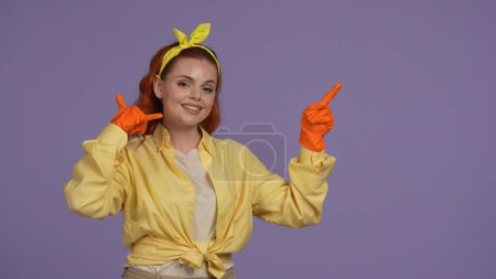 Photo for Everyday cleaning and housekeeping concept. Woman in casual clothing and rubber gloves pointing, shows epty space and calling gesture, smiling face. Isolated on purple background. - Royalty Free Image