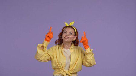 Photo for Everyday cleaning and housekeeping concept. Woman in casual clothing and rubber gloves pointing hands up, smiling at the camera. Isolated on purple background. - Royalty Free Image