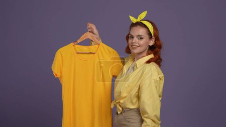 Photo for Everyday cleaning and housekeeping concept. Woman in casual clothing holding trempel with tshirt and smiling at the camera happy face. Isolated on purple background. - Royalty Free Image