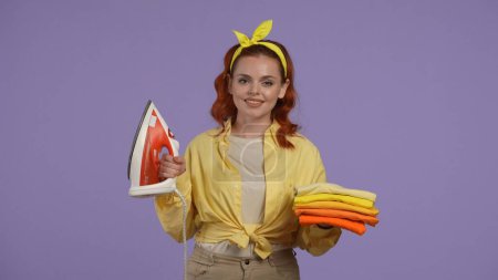 Photo for Everyday cleaning and housekeeping concept. Woman in casual clothing standing holding stack of folded thirts and iron in hands, looking at the camera. Isolated on purple background. - Royalty Free Image