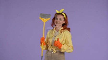 Photo for Everyday cleaning and housekeeping concept. Woman in casual clothing and rubber gloves holding broom showing thumbs up, smiling face, looking at the camera. Isolated on purple background. - Royalty Free Image