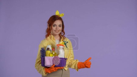 Foto de Concepto diario de limpieza y limpieza. Mujer con ropa casual y guantes de goma sosteniendo cesta con limpiadores, mostrando los pulgares hacia arriba. Aislado sobre fondo púrpura. - Imagen libre de derechos