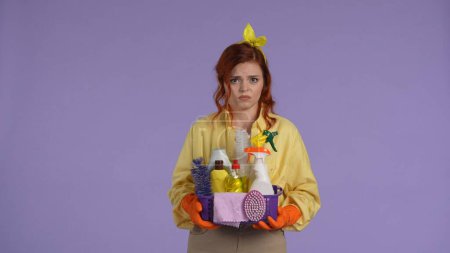 Photo for Everyday cleaning and housekeeping concept. Woman in casual clothing and rubber gloves holding basket with cleaners, worried face, looking at the camera negatively. Isolated on purple background. - Royalty Free Image