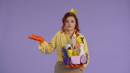Photo for Everyday cleaning and housekeeping concept. Woman in casual clothing and rubber gloves holding basket with cleaners, clueless face expression, shrugs helplessly. Isolated on purple background. - Royalty Free Image